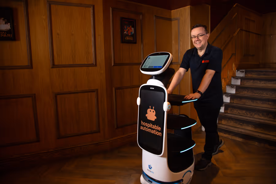 Man standing beside a white autonomous service robot labeled 'hospitable automation' in a wood-paneled room with stairs.