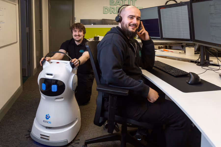 Two men in an office; one sitting at a desk with multiple monitors wearing headphones, the other kneeling behind a white robot with blue eyes labeled Keenon.