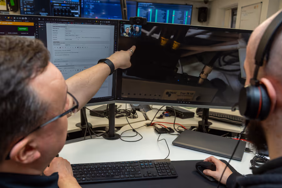 Two men wearing headsets work together at a desk with multiple computer monitors, one man points at the screen while the other uses a mouse.