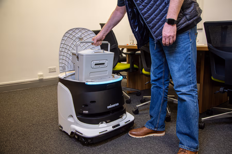 Man placing a metal box into the open top compartment of a white and black OrionStar delivery robot in an office.