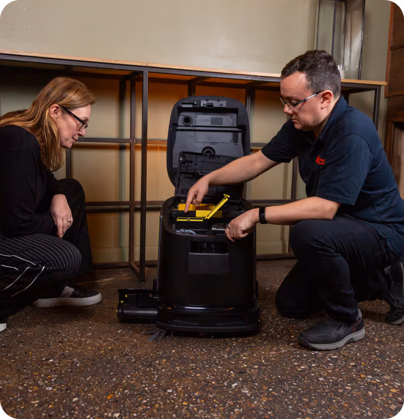 Two people examining the interior components of an open black cleaning robot on a speckled floor indoors.