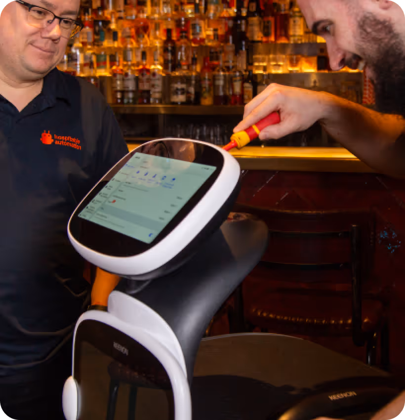 Two men interact with a robot that has a touchscreen interface in a bar setting with bottles on shelves in the background.