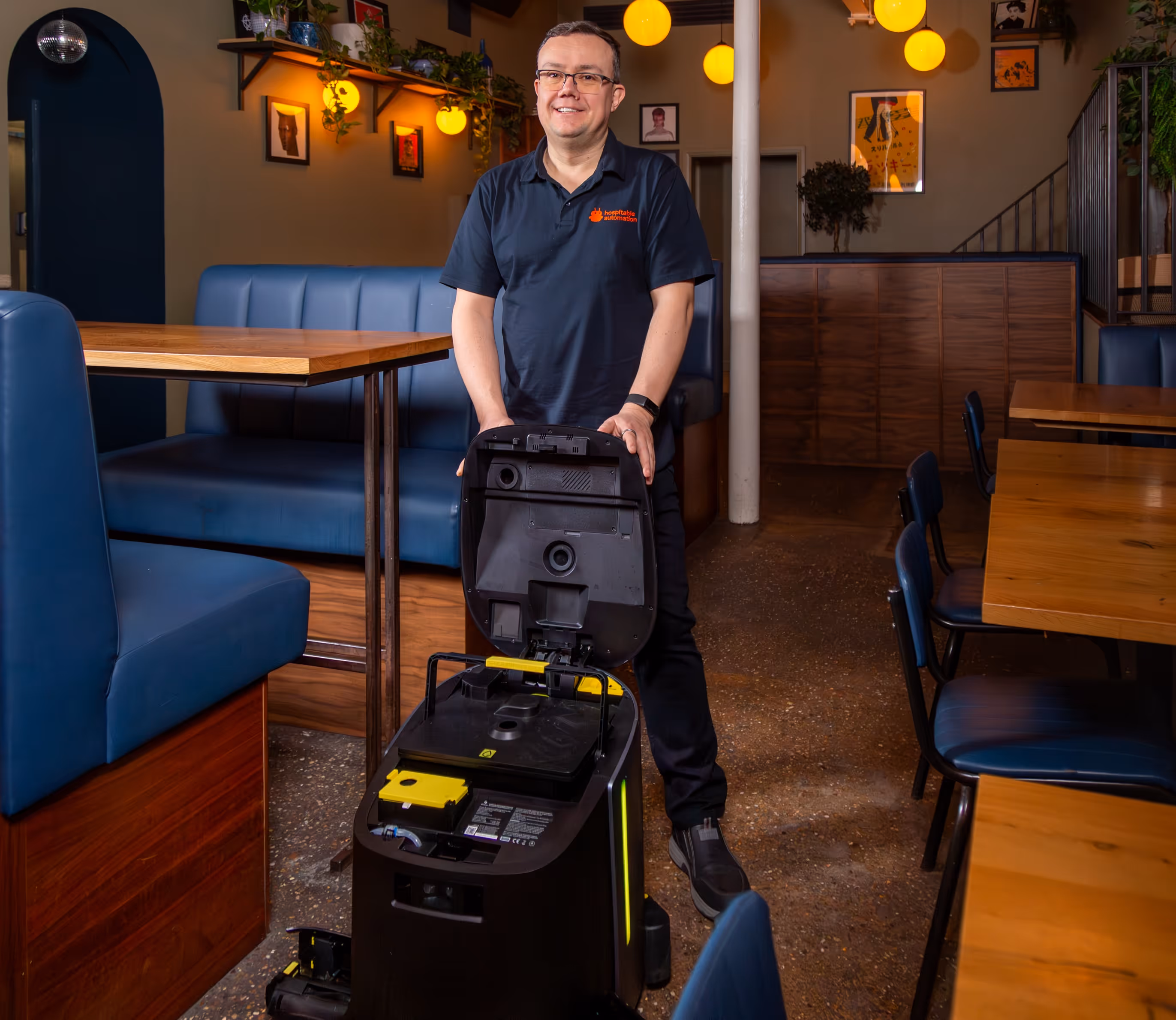 Man in glasses and a navy polo shirt stands in a restaurant holding open a black robotic cleaning machine.
