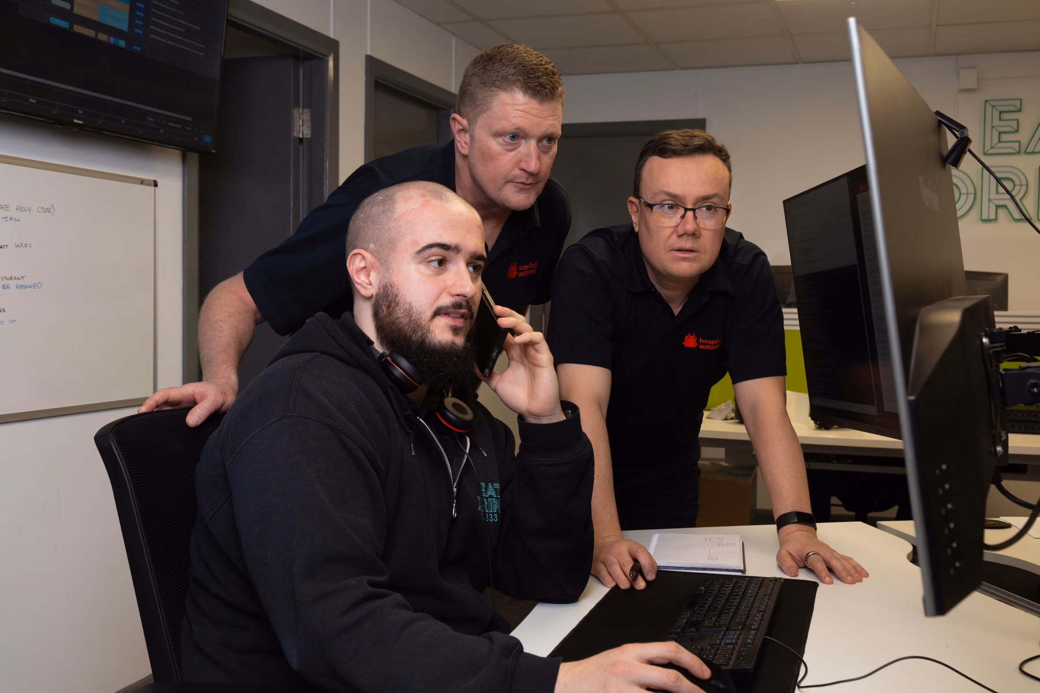 Three men working together at a computer in an office, one is seated and talking on a phone while the others stand looking at the monitor.