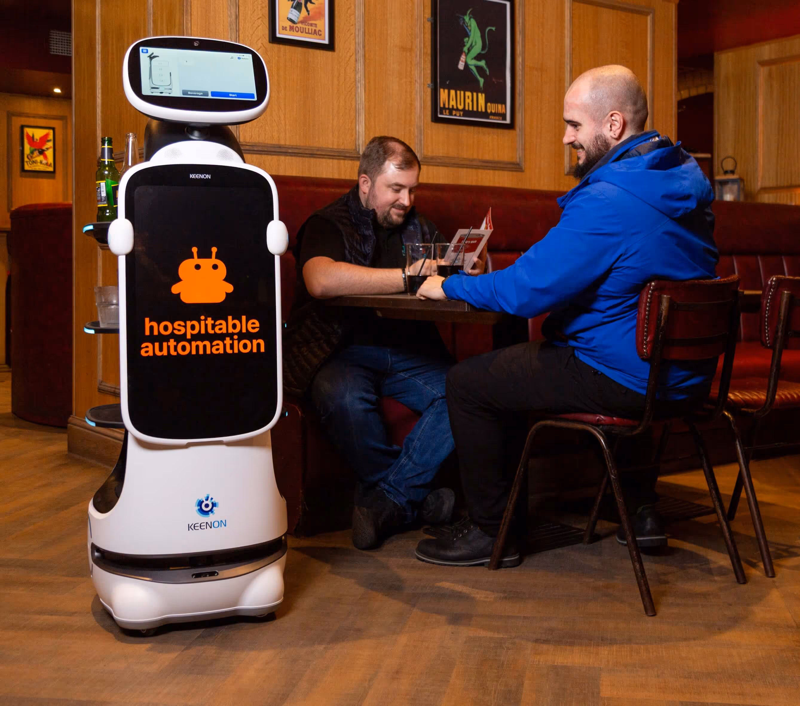 Robot server with 'hospitable automation' text serving drinks to two men seated at a table in a wooden-paneled restaurant.