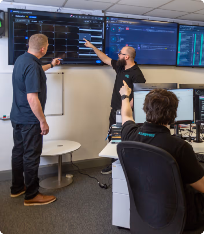Three men in an office looking at and pointing to multiple large monitors displaying calendar and data screens.