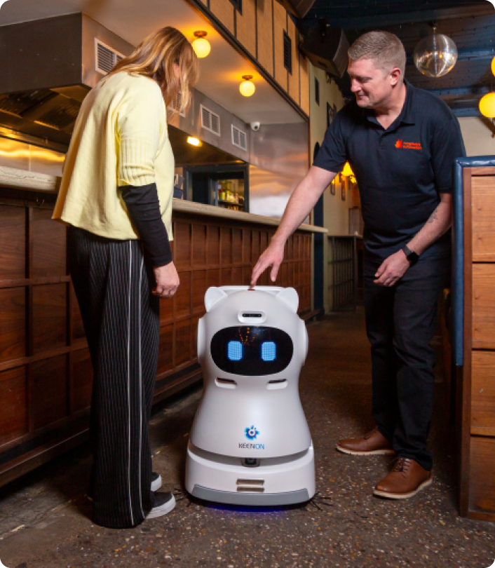 Two people interacting with a white service robot with digital eyes inside a modern restaurant.