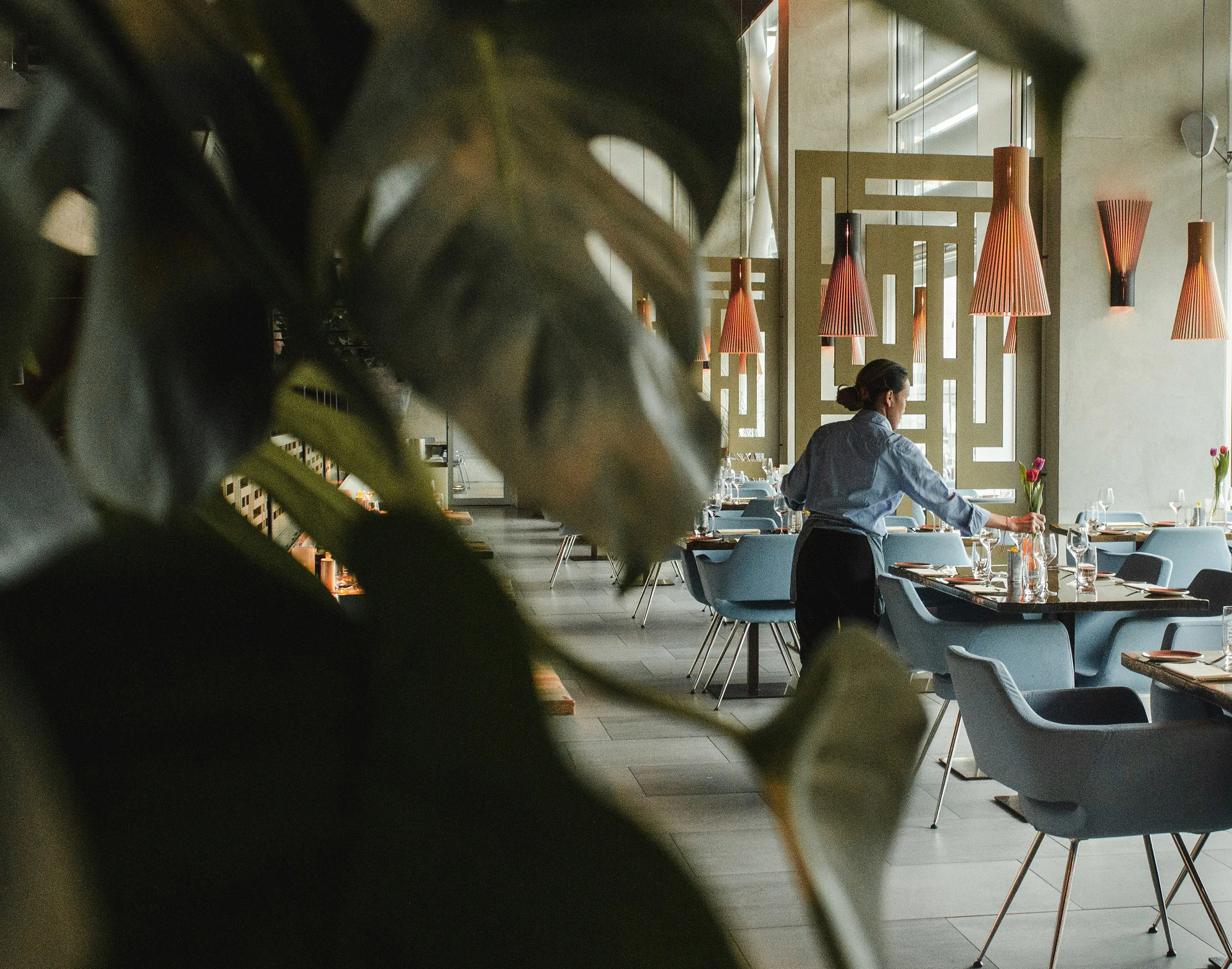 Servitören lägger en blomma i en vas på ett bord i en modern restaurang med blå stolar och pendellampor.