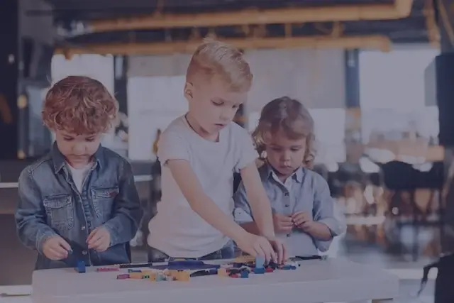 Three young children playing and building with colorful blocks on a white table indoors.