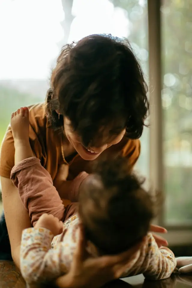 Smiling woman leaning over and holding a baby in a cozy indoor setting with a window in the background.