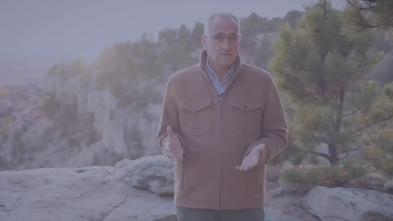 Man wearing glasses and a brown jacket standing outdoors on rocky terrain with trees and cliffs in the background.
