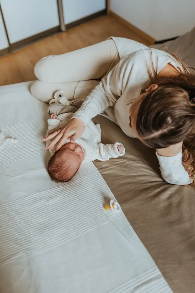 Mother lying on a bed gently touching the forehead of her newborn baby dressed in white.