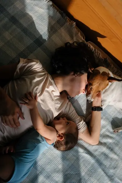 Woman lying on a blanket kissing a dog while a toddler rests on her arm.