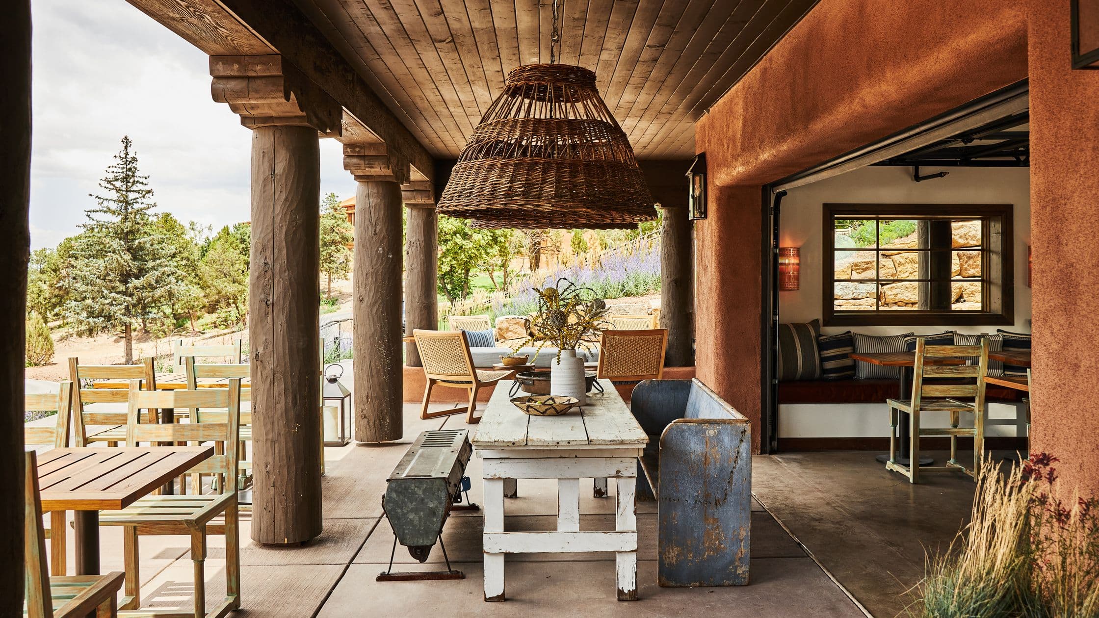 Rustic outdoor patio with wooden pillars, a white wooden table, metal bench, wicker chairs, and large woven pendant lights.
