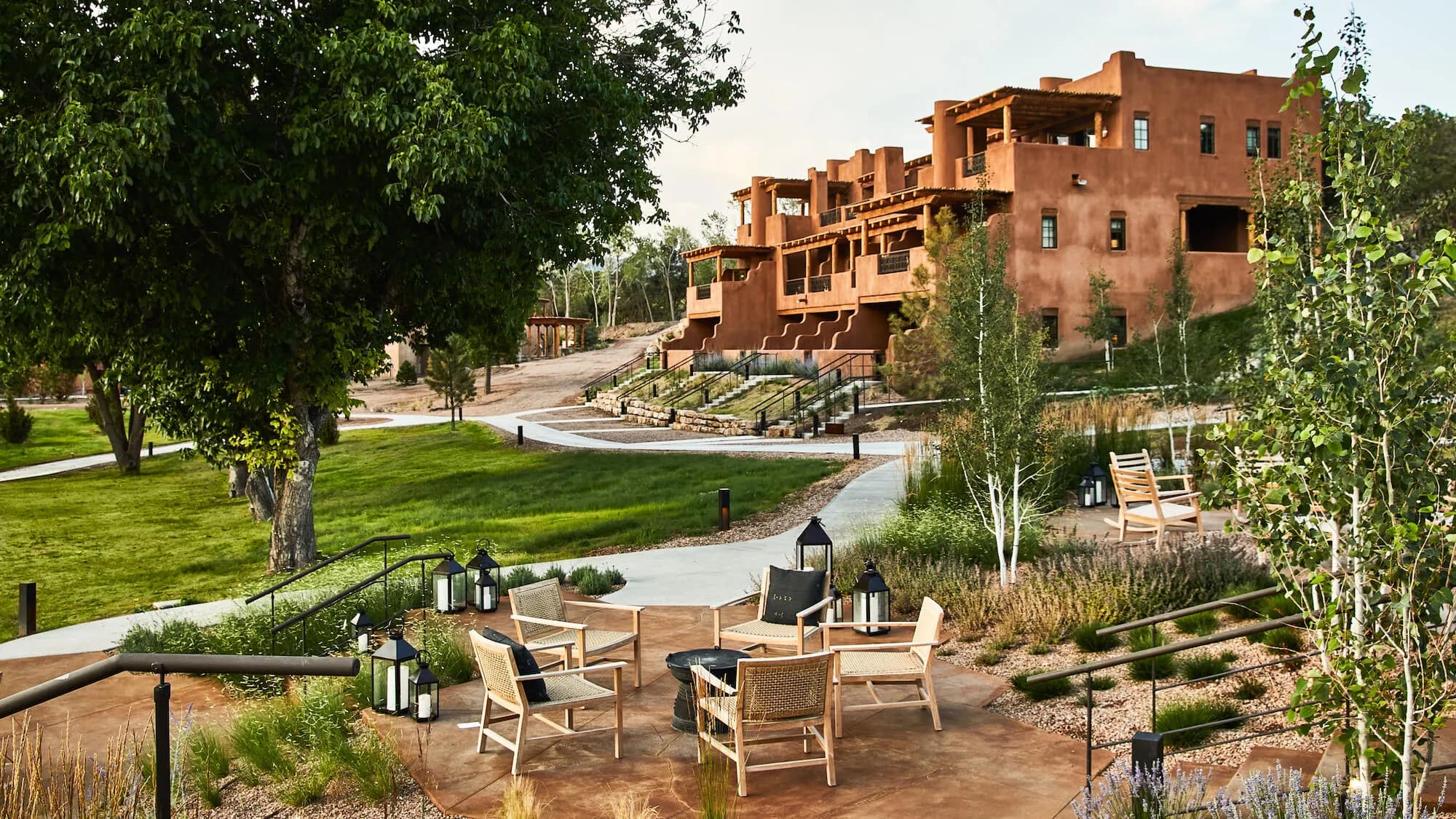 Outdoor seating area with six wooden chairs arranged around a fire pit, surrounded by greenery and paths, with a large adobe-style building in the background.