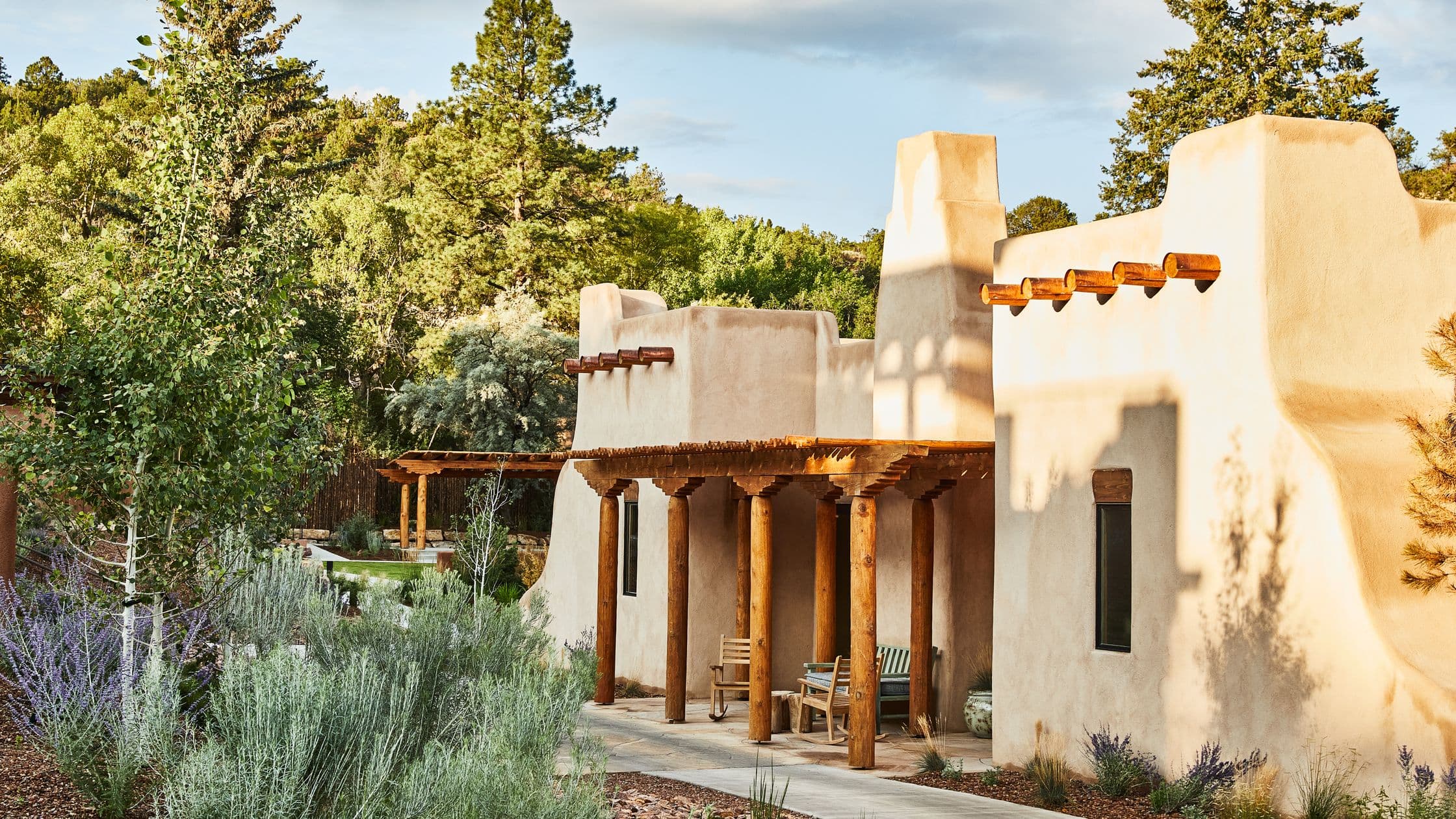 Adobe-style house with wooden columns and pergola surrounded by lush greenery and trees under a partly cloudy sky.