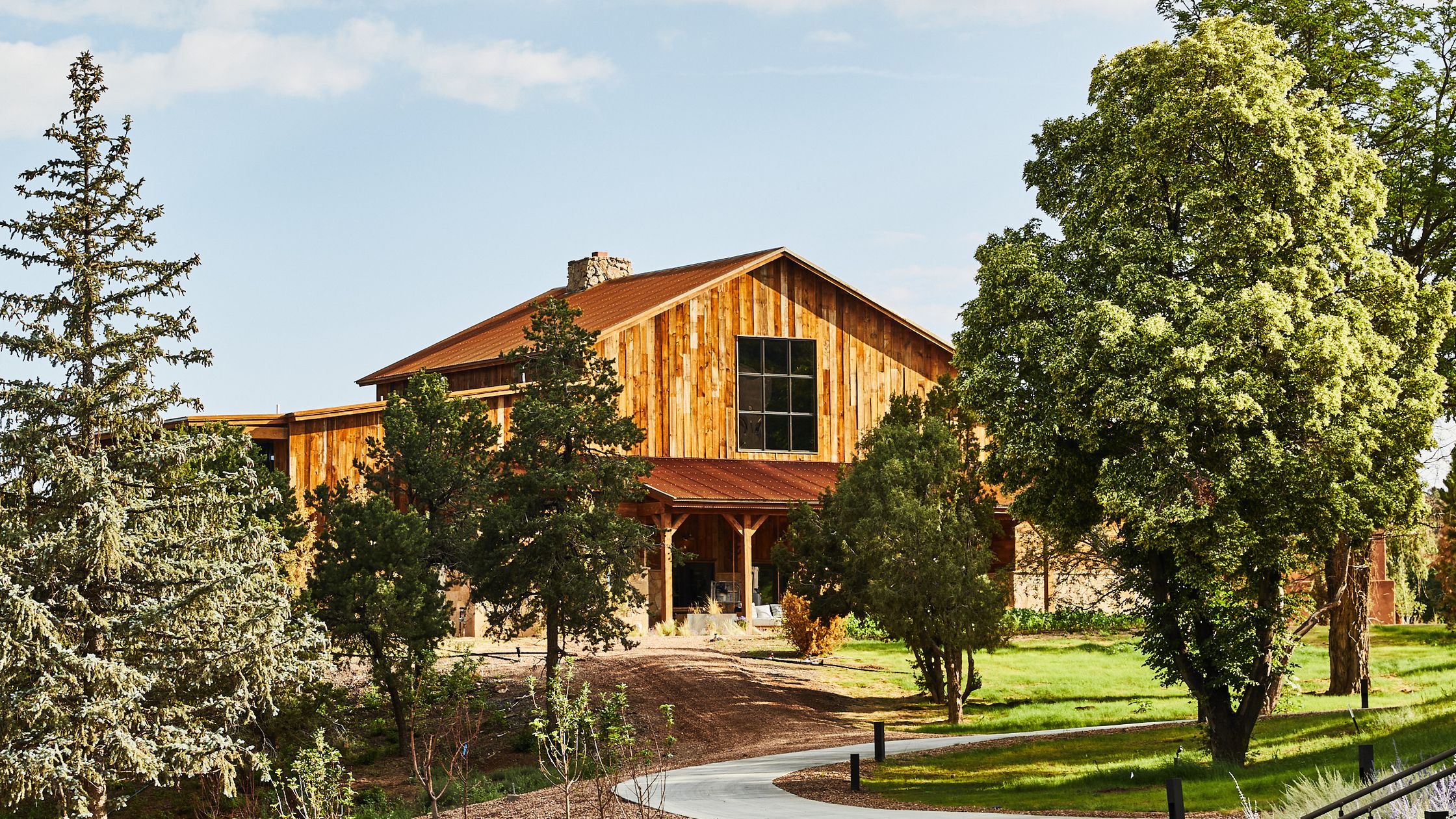 Large wooden barn-style building with a rust-colored roof surrounded by various green trees and a curved concrete path.
