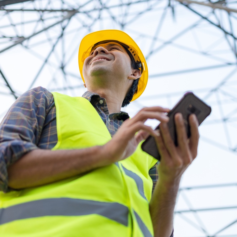Utility worker in safety vest using a tablet near power lines.