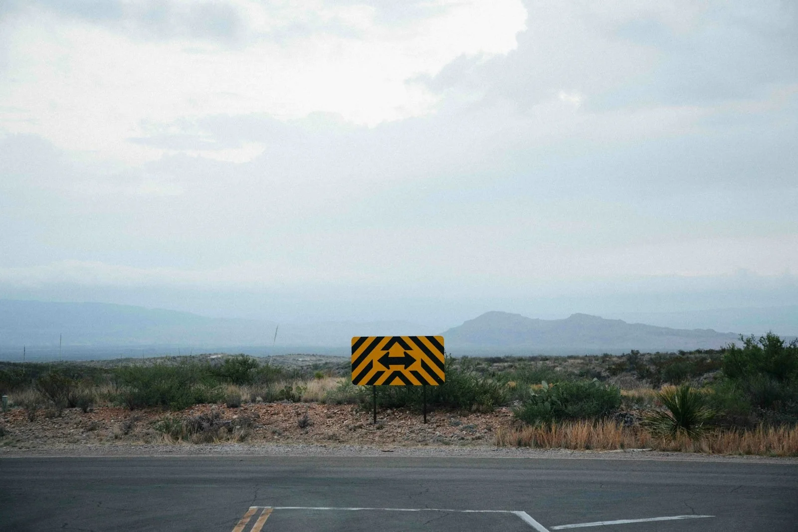 A yellow and black striped hazard sign with a double-headed horizontal arrow stands in a desert landscape under an overcast sky, positioned beyond a paved road