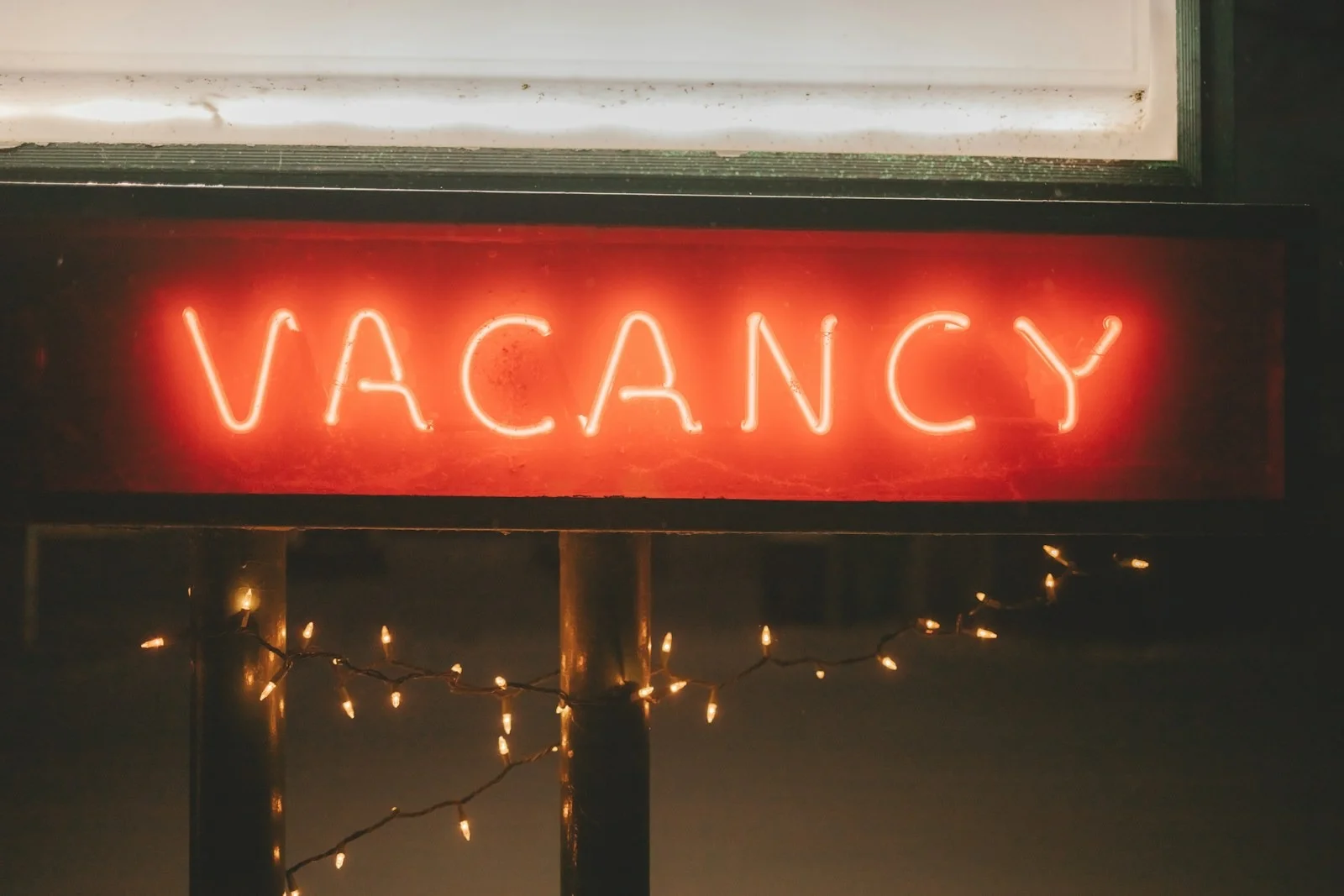A glowing red neon sign displays the word "VACANCY" against a dark background, with small warm-toned fairy lights draped underneath
