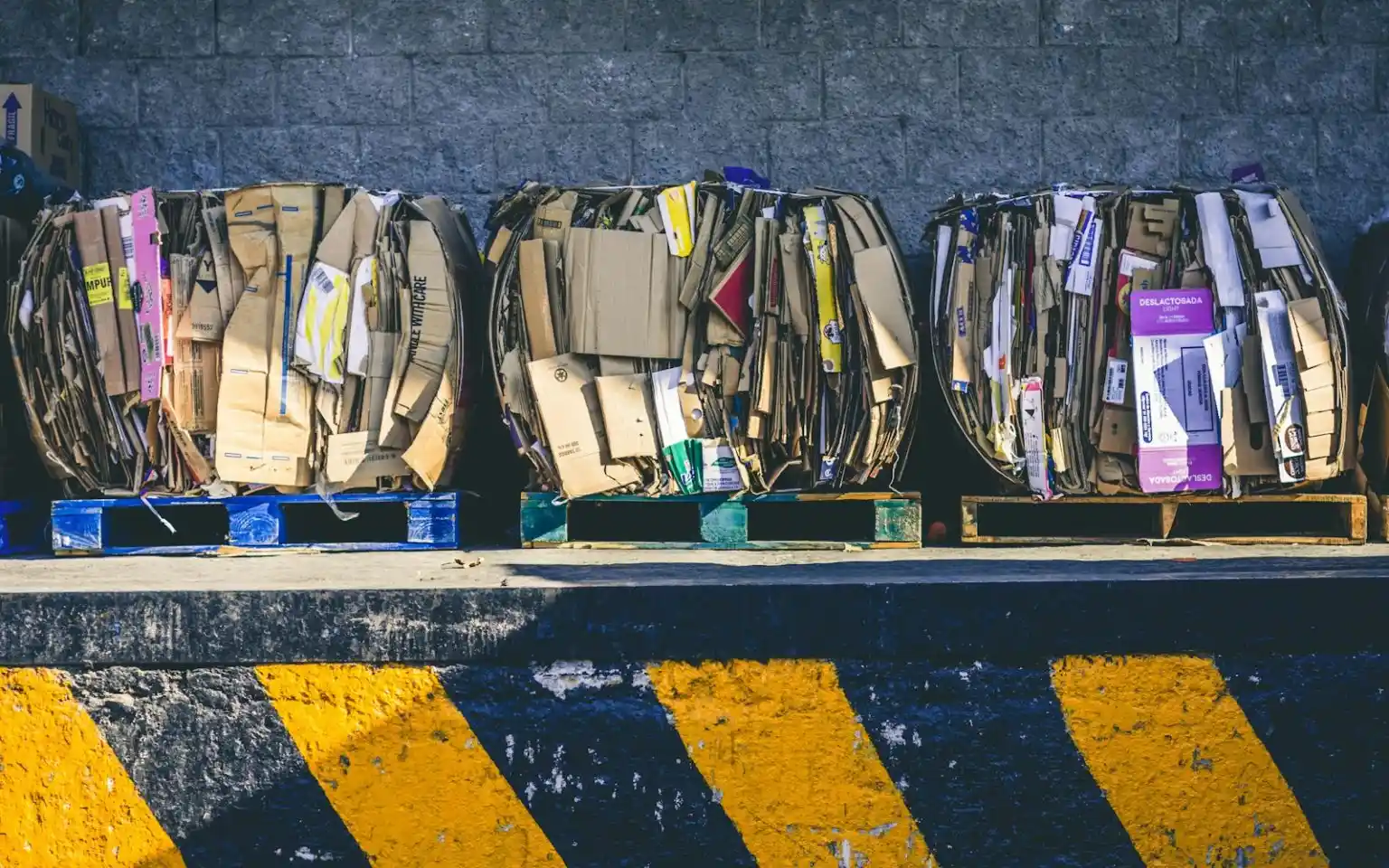 Three large, round bales of crushed cardboard sit on blue and green wooden pallets along a loading dock with a yellow and black striped safety edge