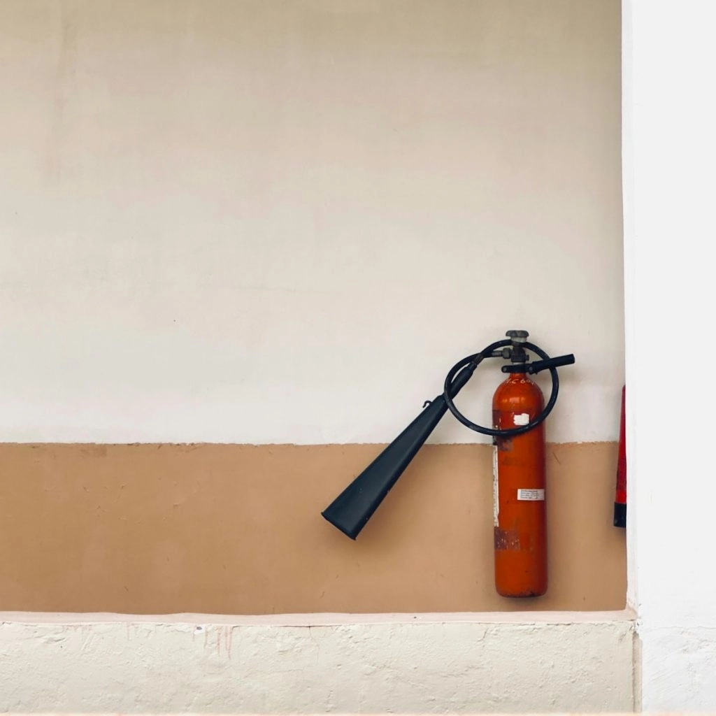 An orange fire extinguisher with a long black nozzle is mounted on a two-toned wall, with the top half being white and the bottom half a light tan