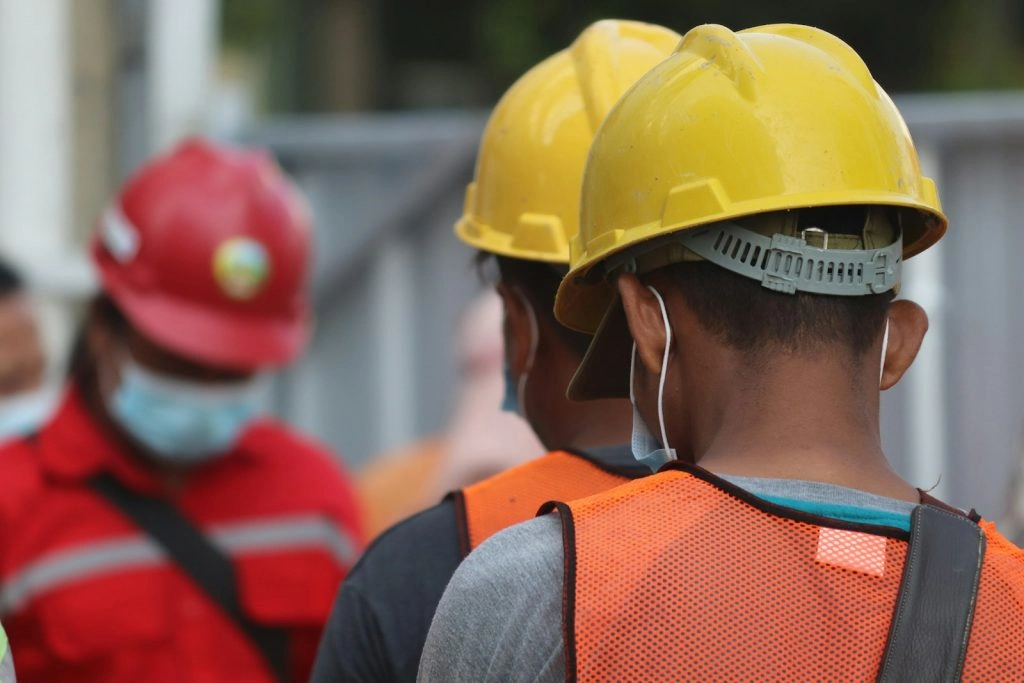 Rear view of two workers wearing yellow hard hats and high-visibility orange vests, looking towards a supervisor in a red uniform and face mask