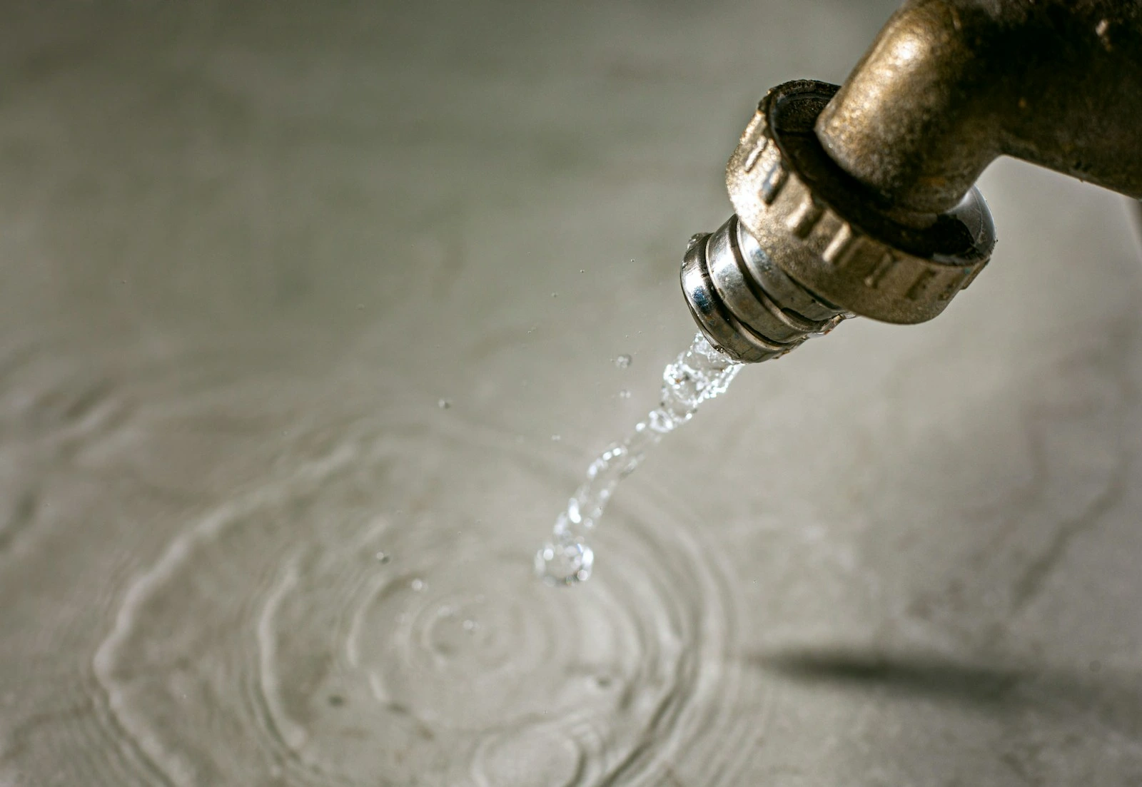Close-up of a brass outdoor faucet dripping water into a shallow pool, creating concentric ripples on the surface