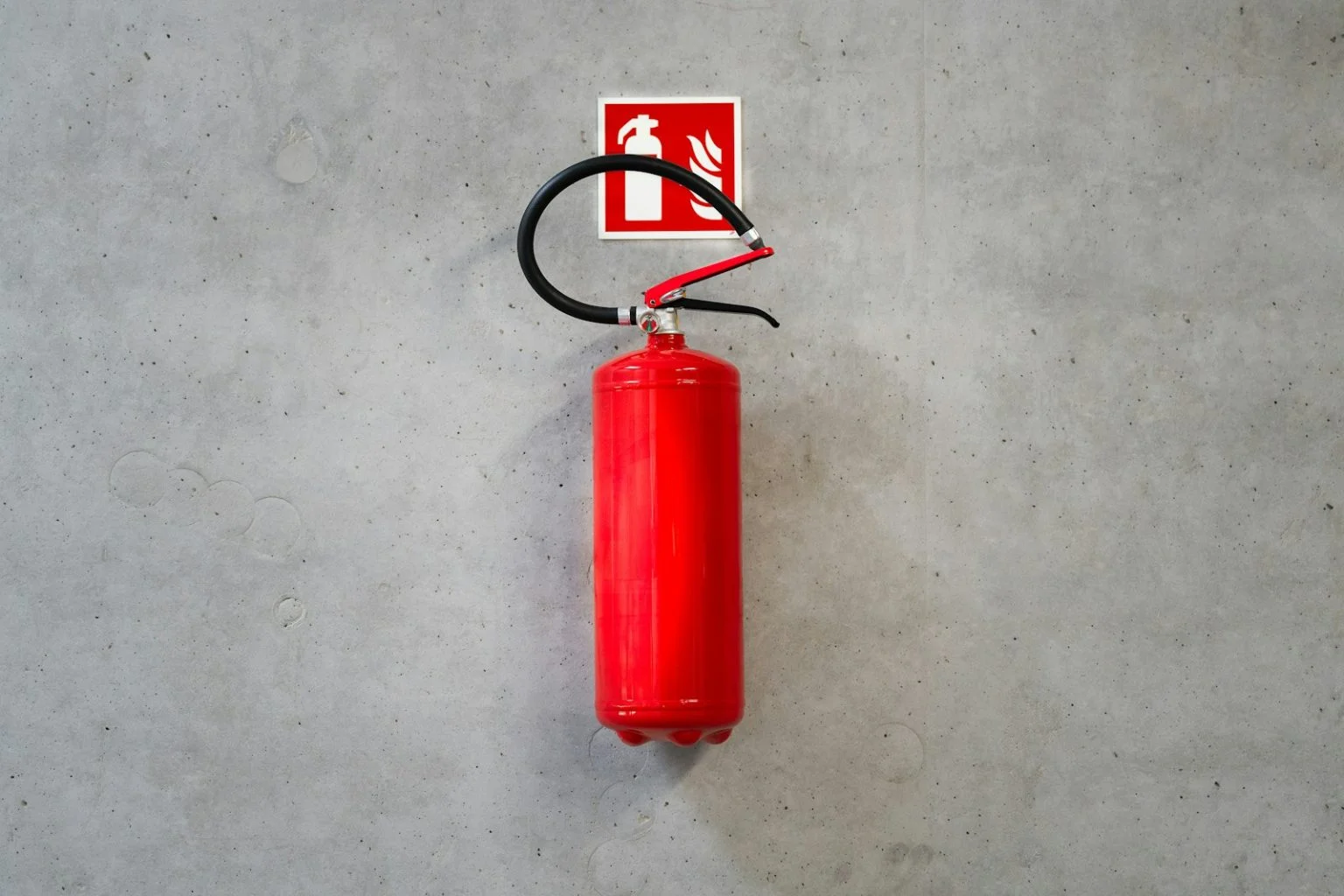 A bright red fire extinguisher is mounted on a smooth, gray concrete wall, positioned directly below a square red and white safety sign depicting a fire extinguisher and flames