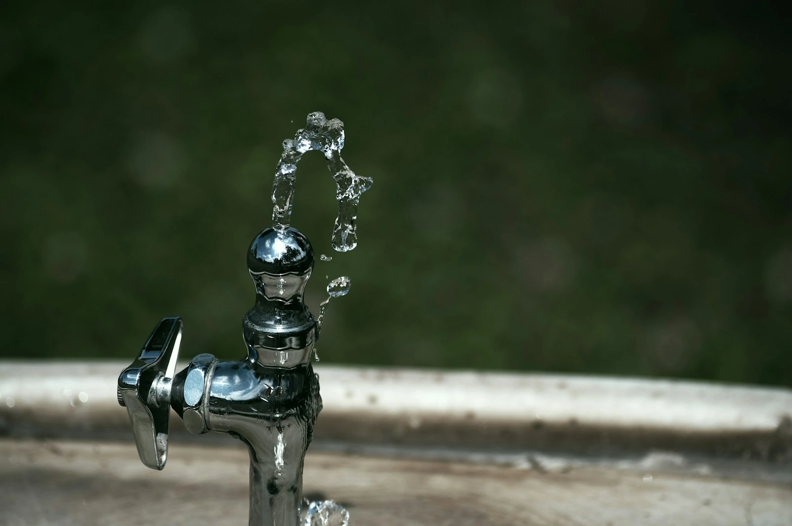 A metal drinking fountain outdoors with a small stream of water arching upwards against a blurred dark green background