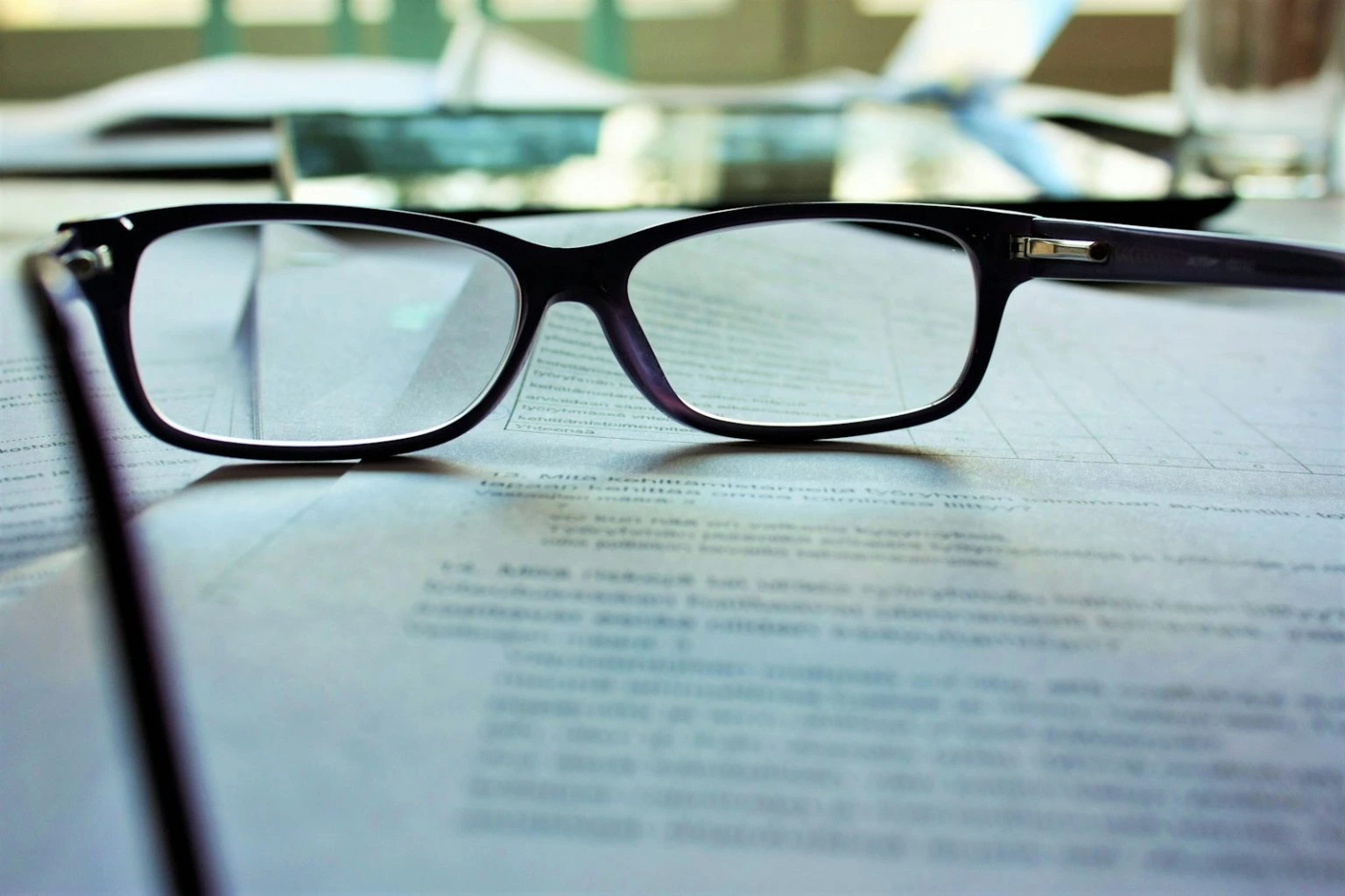 A close-up shot of black-framed eyeglasses resting on top of printed documents, with the background softly blurred to focus on the lenses and the text underneath