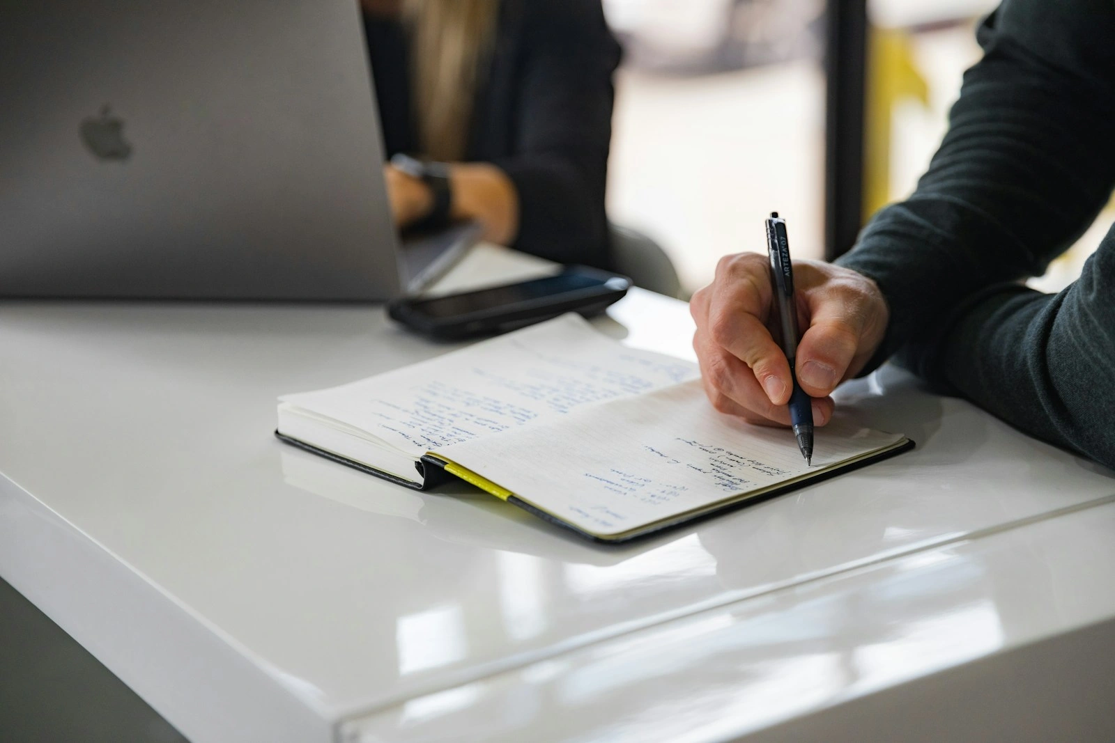 Close-up of a person's hand writing in a notebook with a pen on a white desk next to a laptop
