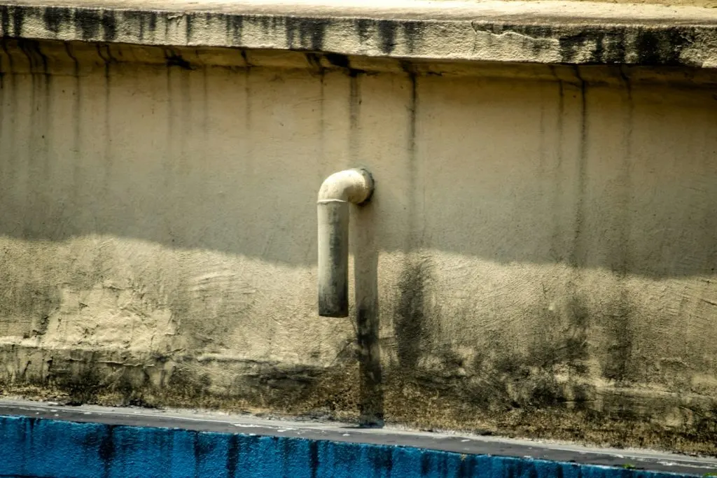 A weathered, beige concrete wall features a single L-shaped pipe protruding and pointing downward, with dark water stains streaking the surface below it