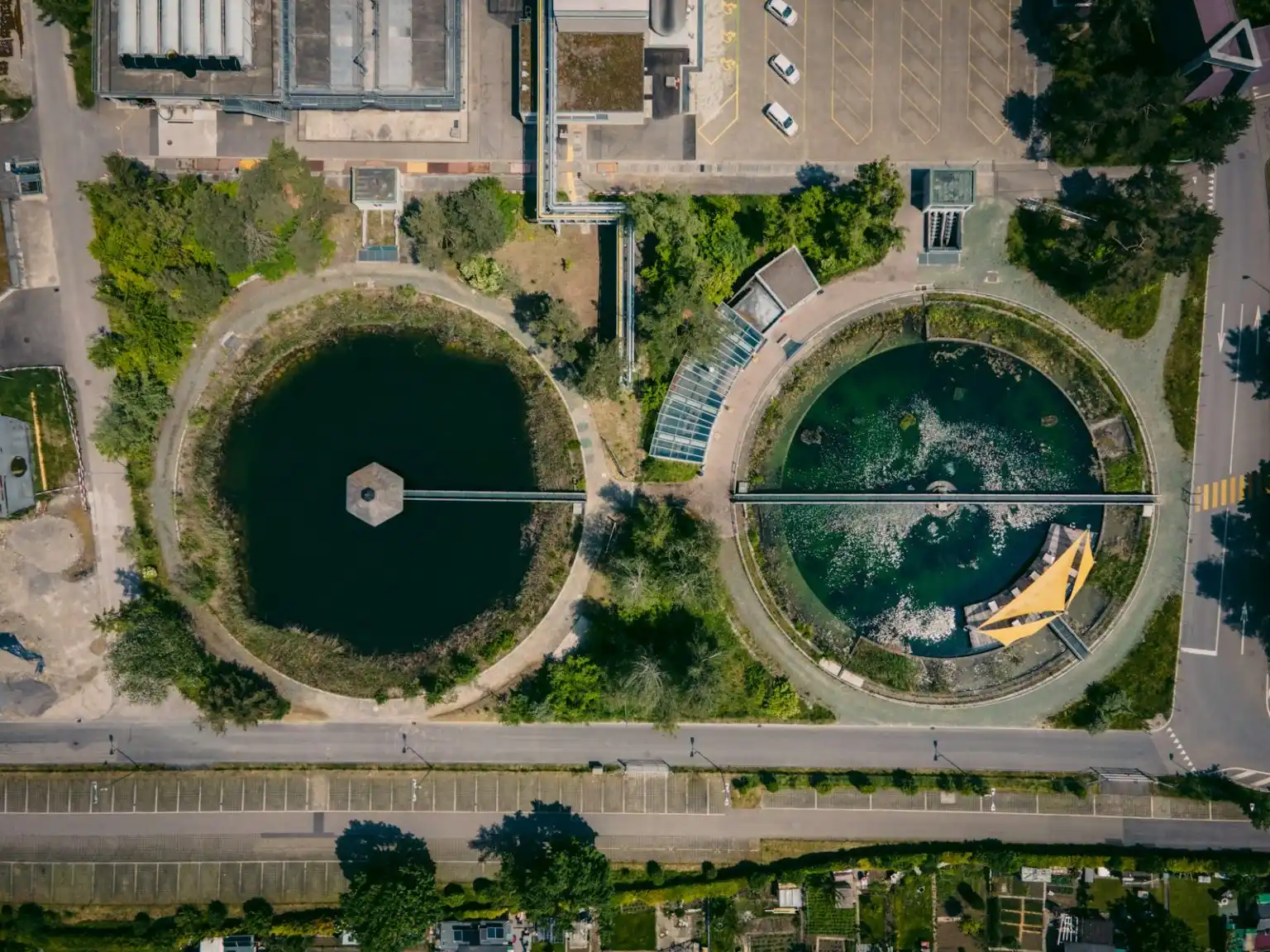 An aerial, top-down view of a water treatment facility featuring two large, circular clarification tanks surrounded by green trees, access roads, and industrial buildings