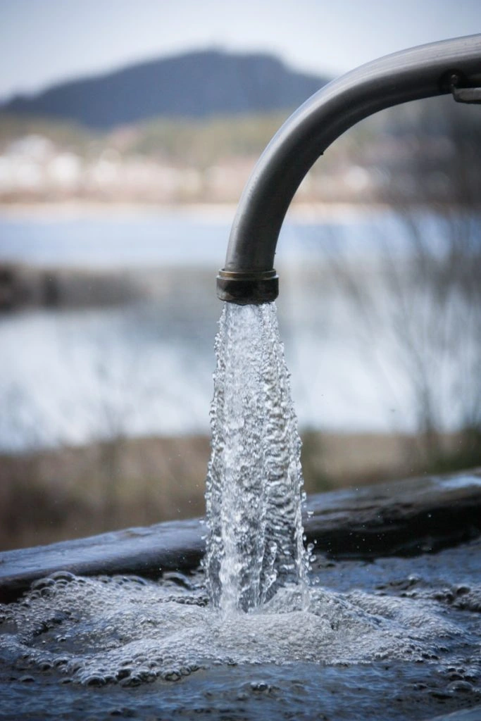 A steady stream of clear water pouring from a metal outdoor faucet into a bubbling stone basin, with a blurred natural landscape and a mountain in the background