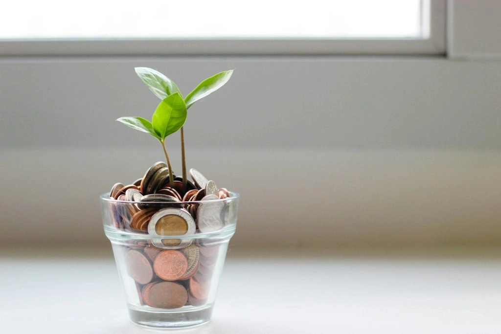 A small green plant growing out of a glass jar filled with various coins, set against a bright, blurred background