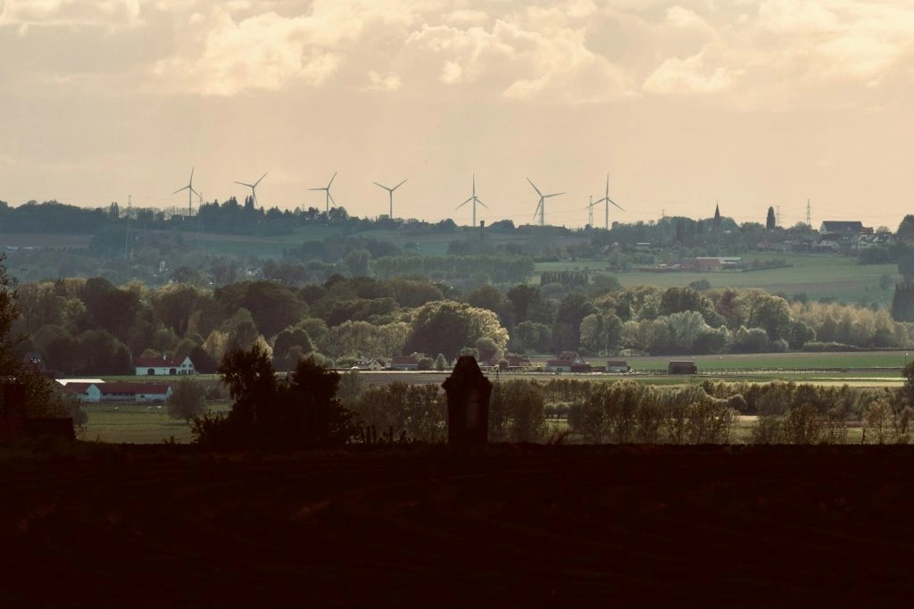 A line of white wind turbines situated on a distant hill above a green countryside with houses and trees under a cloudy sky