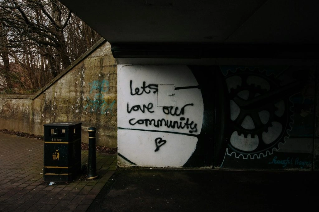 Graffiti on an underpass wall with the handwritten message, "lets love our community" next to a small heart symbol and a public trash bin