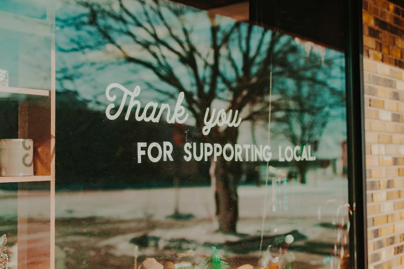 A shop window with the white text "Thank you FOR SUPPORTING LOCAL." reflecting bare trees and a street scene