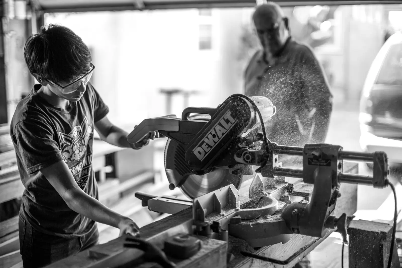 A black and white photo of a person wearing safety glasses and using a DeWalt miter saw to cut wood in a workshop, with another person watching in the background