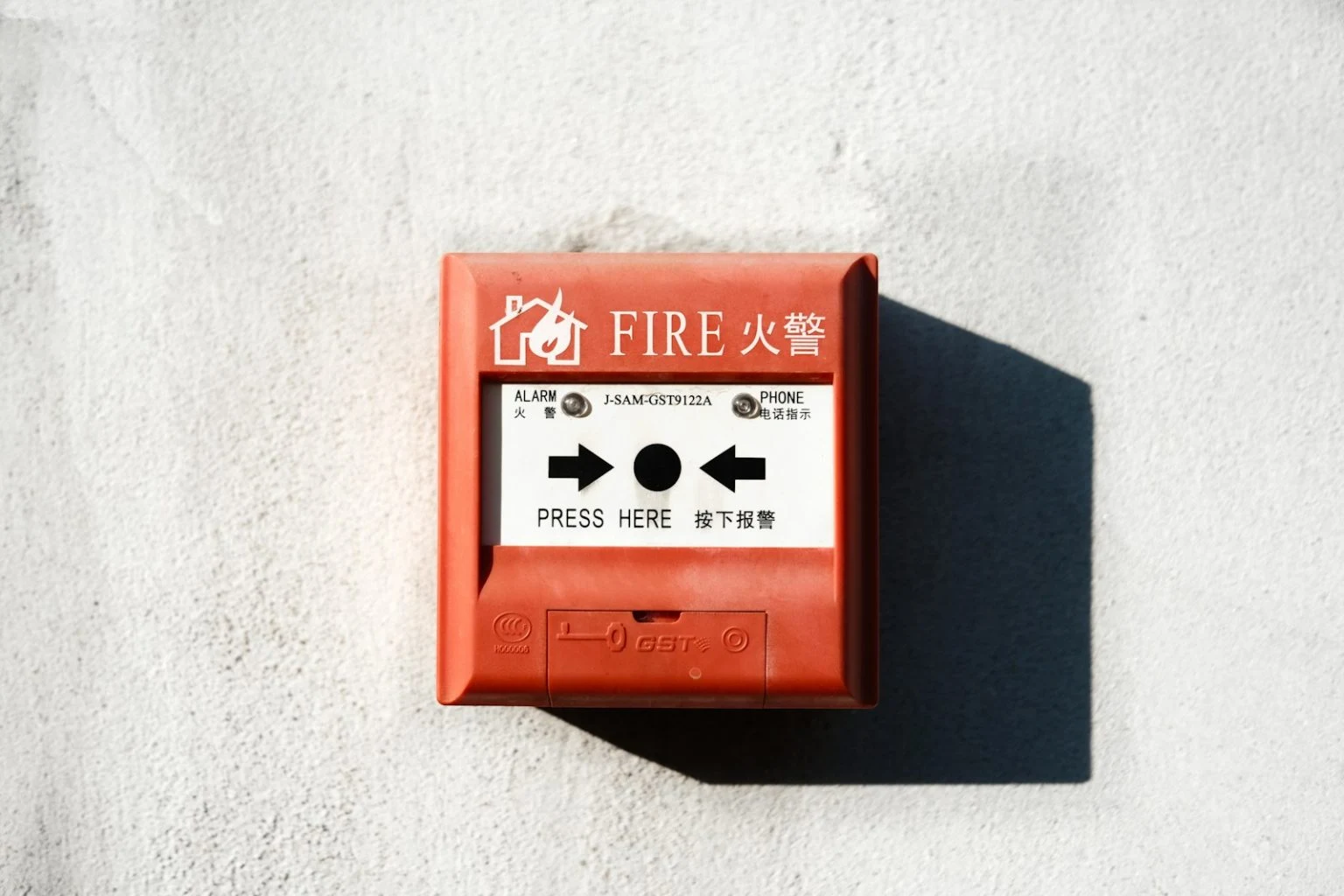 A red square fire alarm manual call point is mounted on a textured white wall, with "FIRE" written in English and Chinese, and instructions saying "PRESS HERE" alongside arrows pointing toward a central button