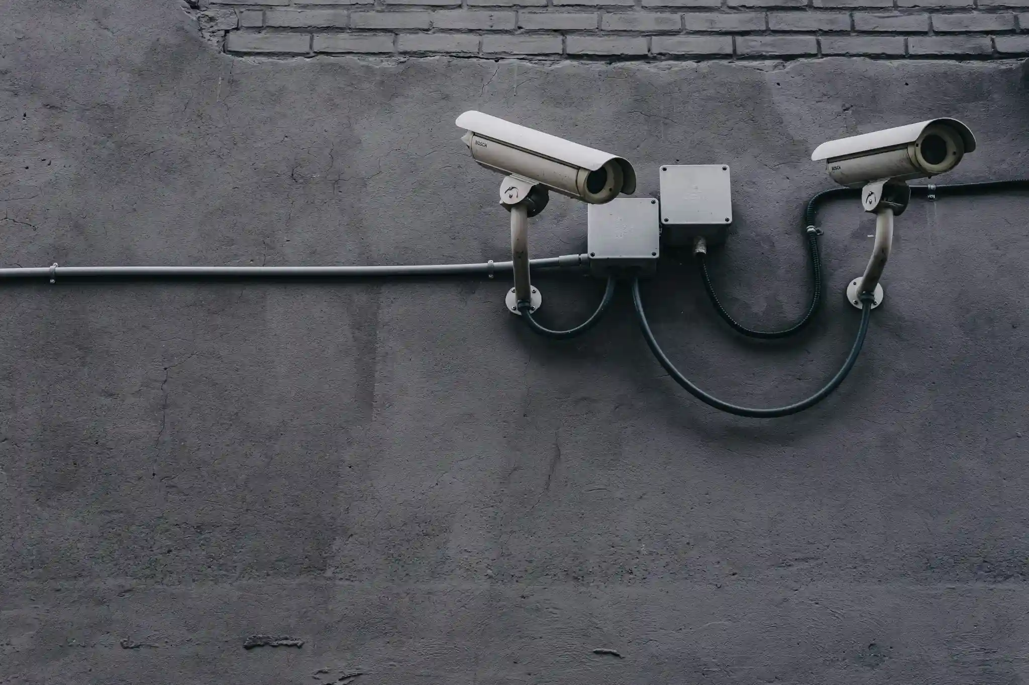 Two white Bosch security cameras are mounted on a gray concrete wall, connected by wires to two central junction boxes