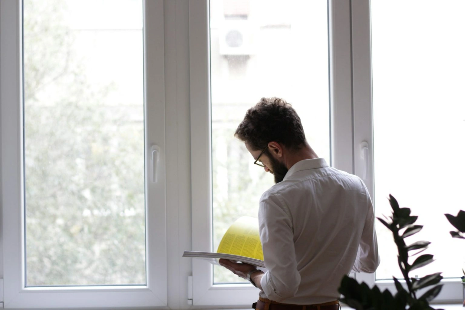 Seen from behind, a man with a beard and glasses, wearing a white shirt, stands in front of a bright window while reading a magazine with yellow pages