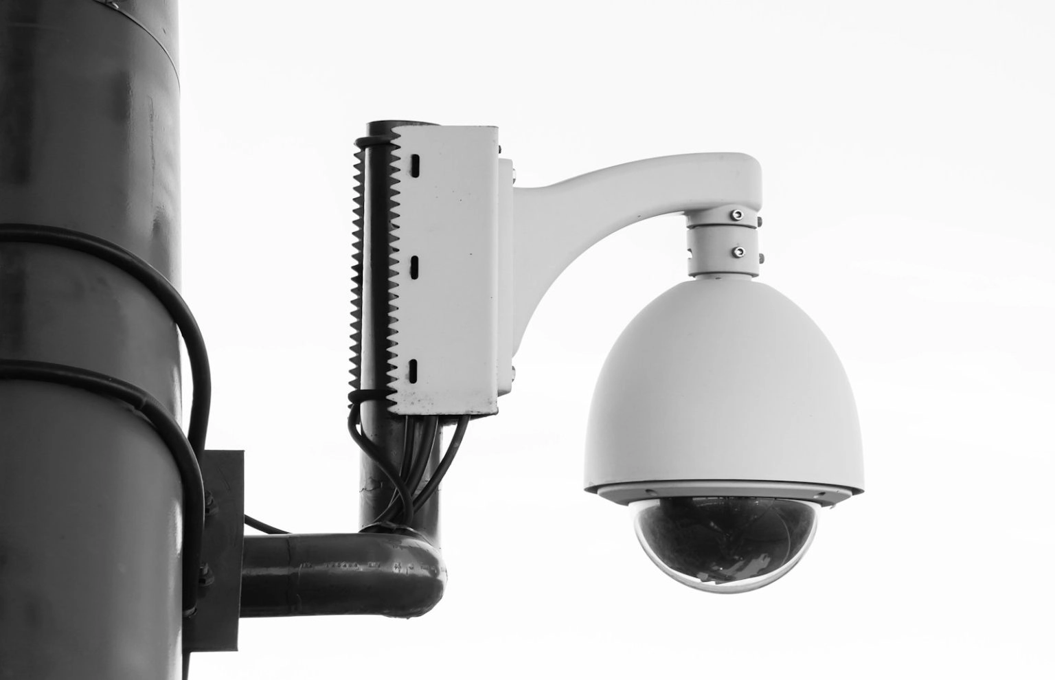 A white dome-shaped security camera is mounted to a thick metal pole with a curved bracket against a plain white background
