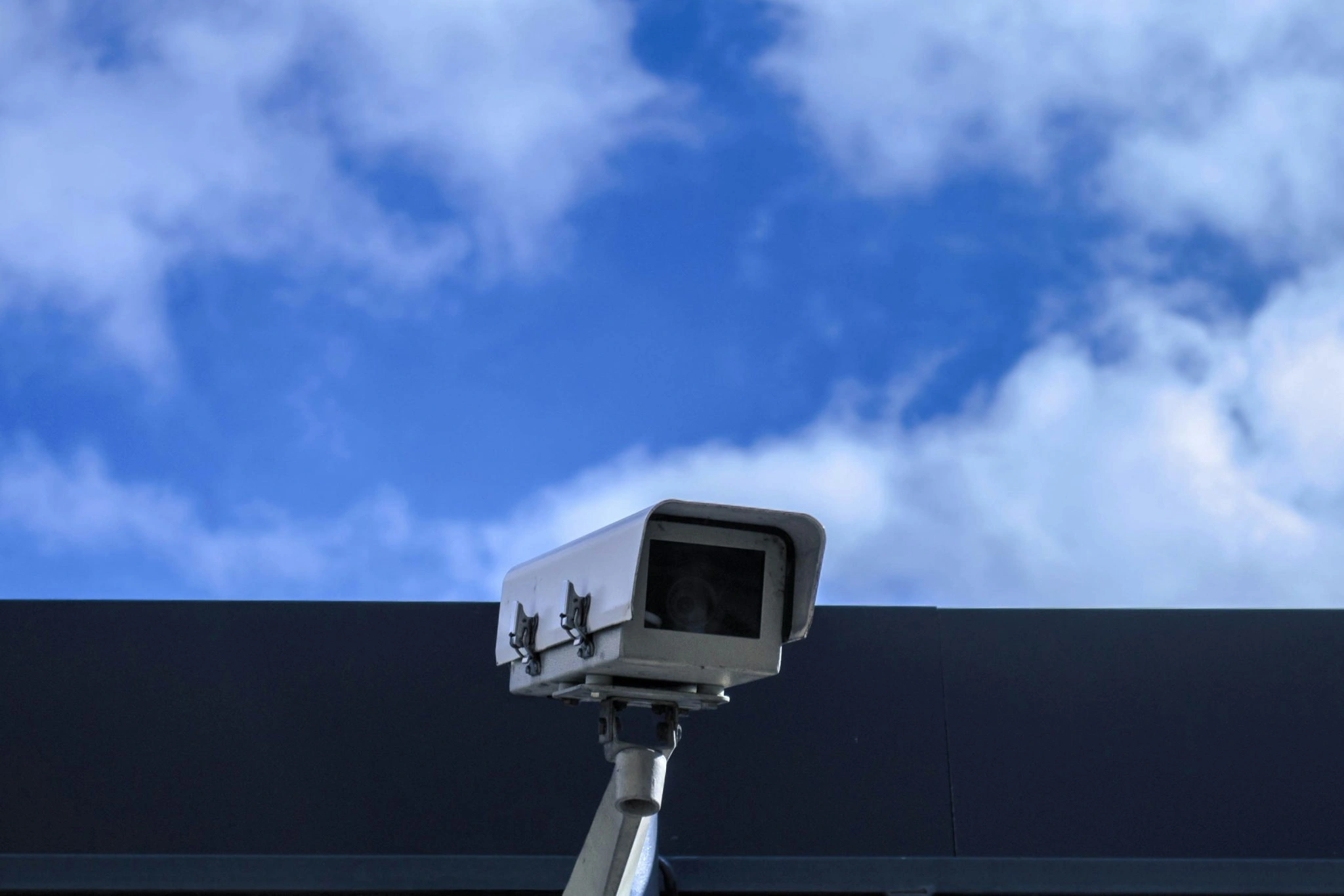 A white rectangular security camera is mounted onto a dark ledge, angled downward against a bright blue sky with wispy white clouds