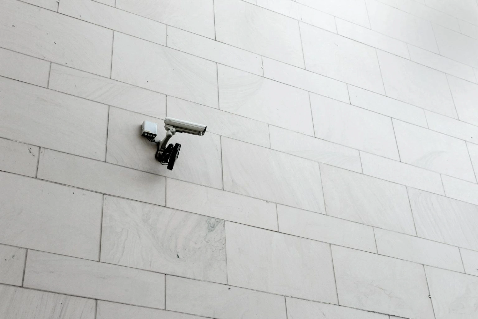 A white security camera is mounted on a light-colored, tiled stone wall and pointed to the right