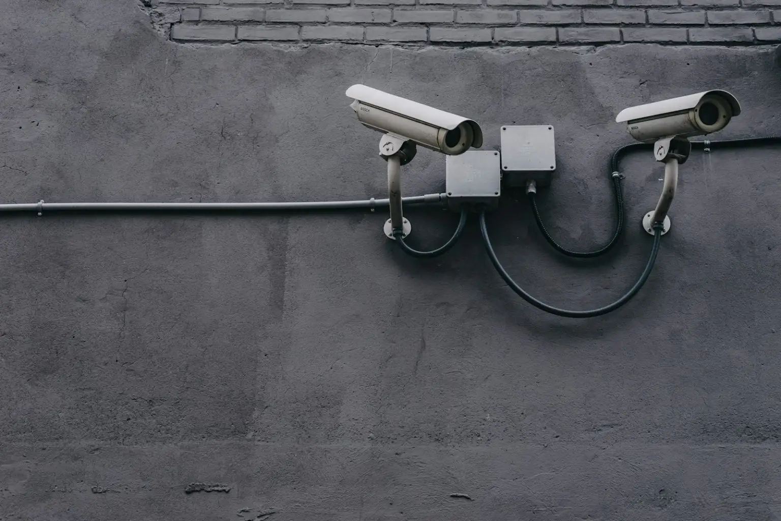 Two white security cameras are mounted on a gray concrete wall, connected by wires to two central junction boxes