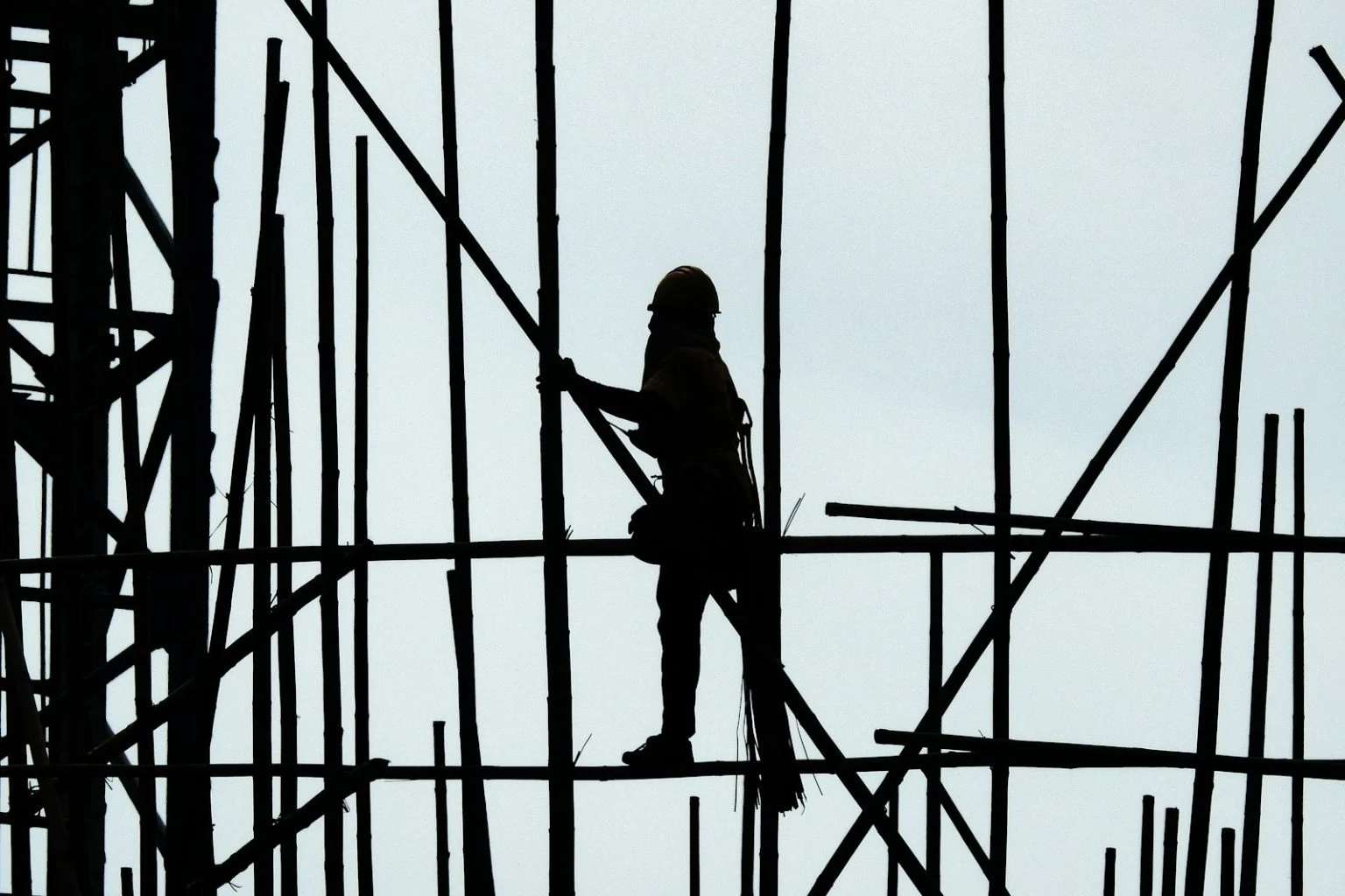 A silhouette of a construction worker wearing a hard hat, standing on a scaffolding structure composed of many vertical and horizontal poles against a bright, hazy sky