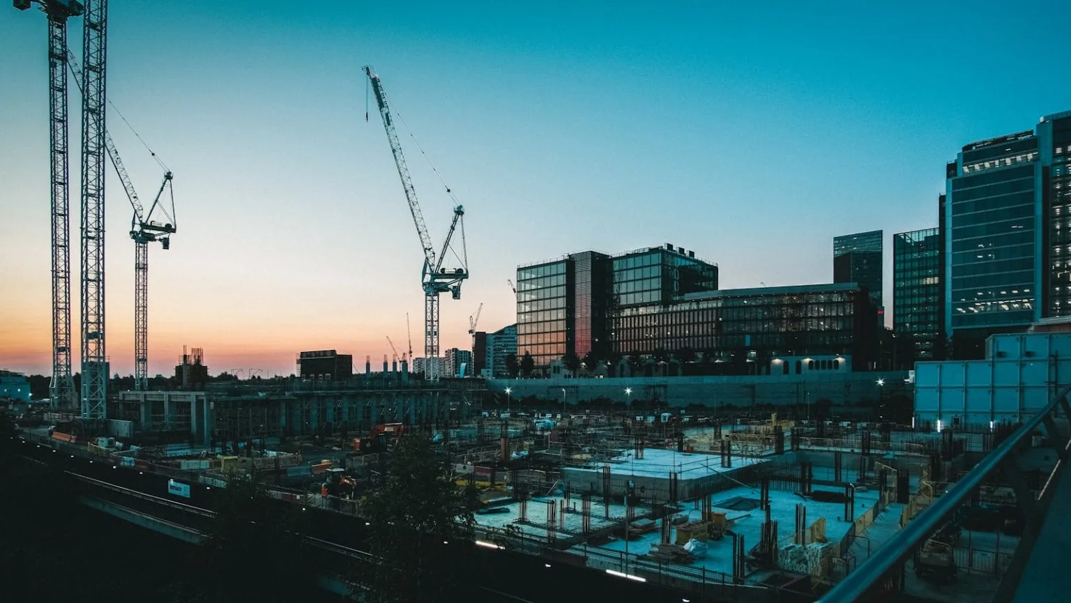 A wide shot of a large construction site at dusk, featuring several tall cranes and unfinished building foundations in the foreground, with modern glass office buildings illuminated in the background under a gradient sunset sky