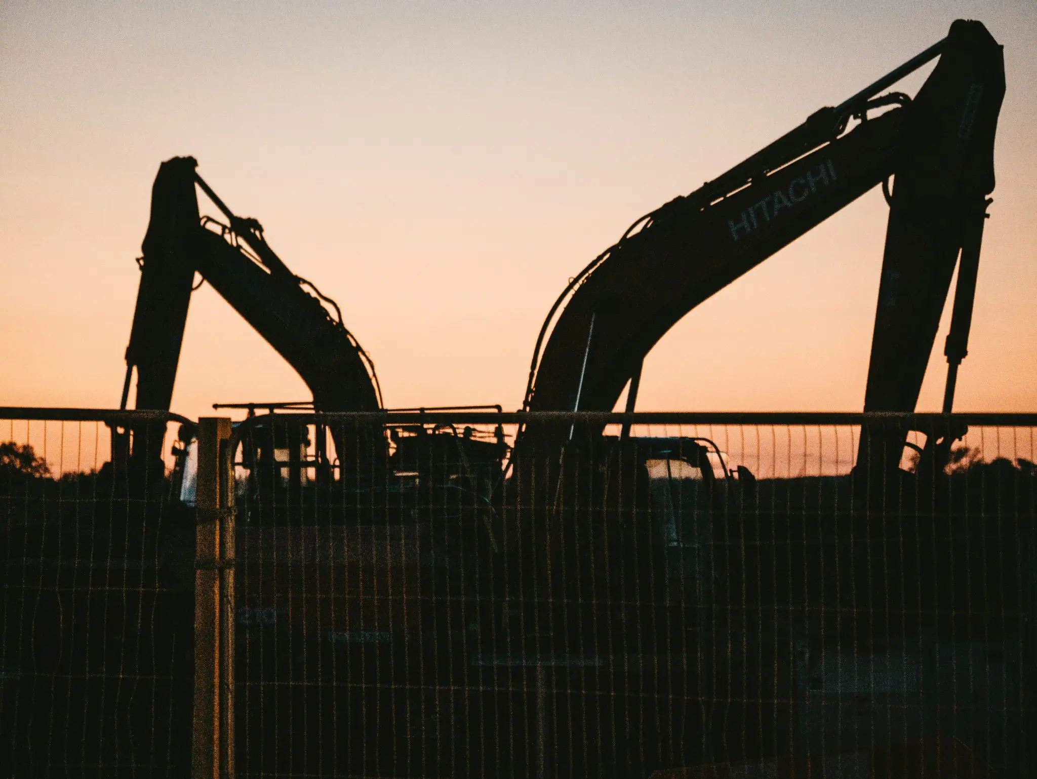 The silhouette of two large Hitachi excavators behind a wire fence against an orange and yellow sunset sky